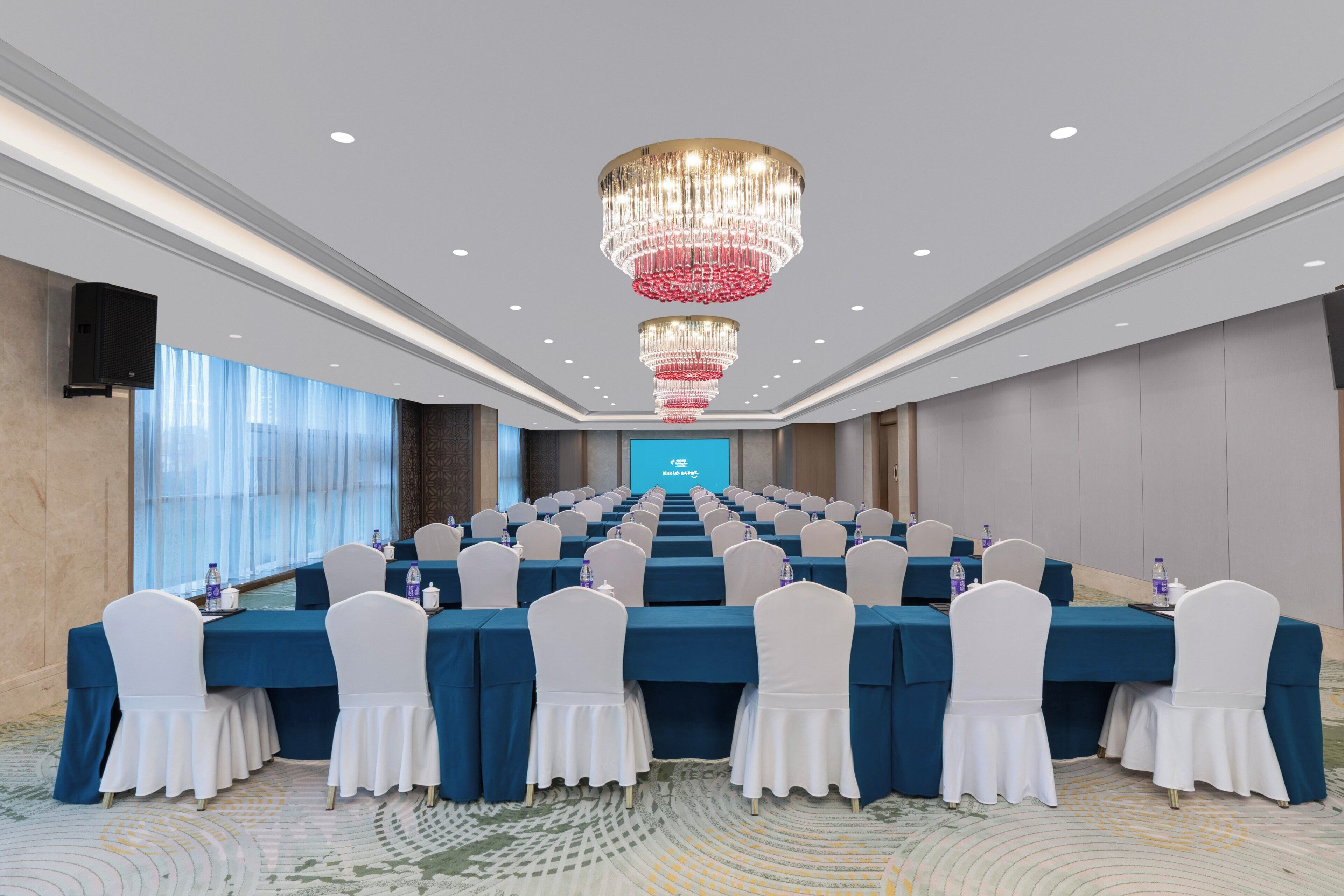 A conference room with rows of tables, white chairs, and a large screen at the front.
