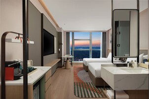 Habitación de hotel con cama, escritorio, televisión, baño y vista a la ciudad al atardecer.