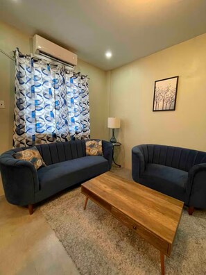 Ein Wohnzimmer mit zwei Sofas, einem Couchtisch, einem gemusterten Vorhang, einer Wandlampe und einer Klimaanlage.