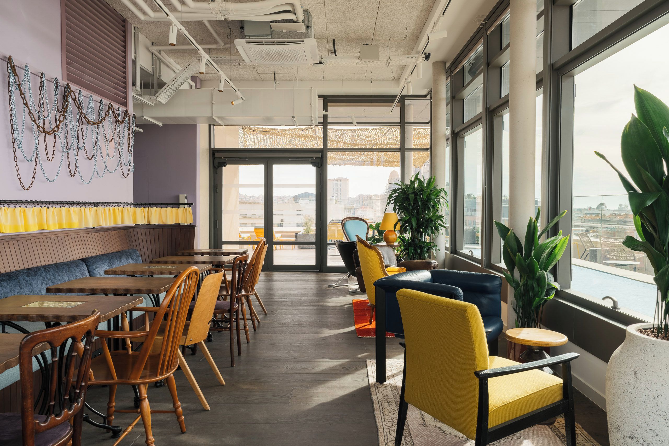 Ein modernes Restaurant mit hoher Decke, freiliegenden Lüftungsrohren und Blick auf die Stadt durch große Fenster.