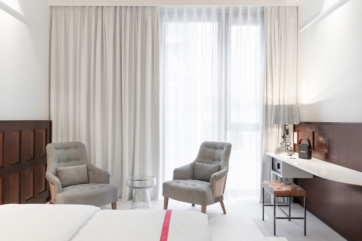 Une chambre d’hôtel comprenant un lit, deux fauteuils, une petite table, un bureau avec une lampe et une grande fenêtre avec des rideaux.