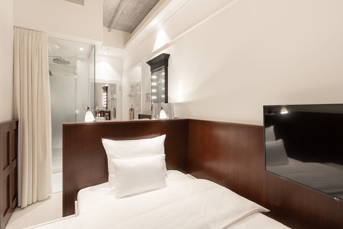 Une chambre d’hôtel équipée d’un lit, d’un grand miroir, d’un téléviseur à écran plat et d’une salle de bain visible à travers une porte en verre.
