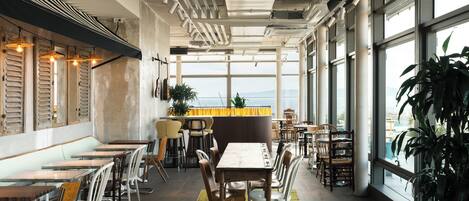 Interior de un restaurante con techos altos, conductos expuestos y ventanas amplias que ofrecen una vista.