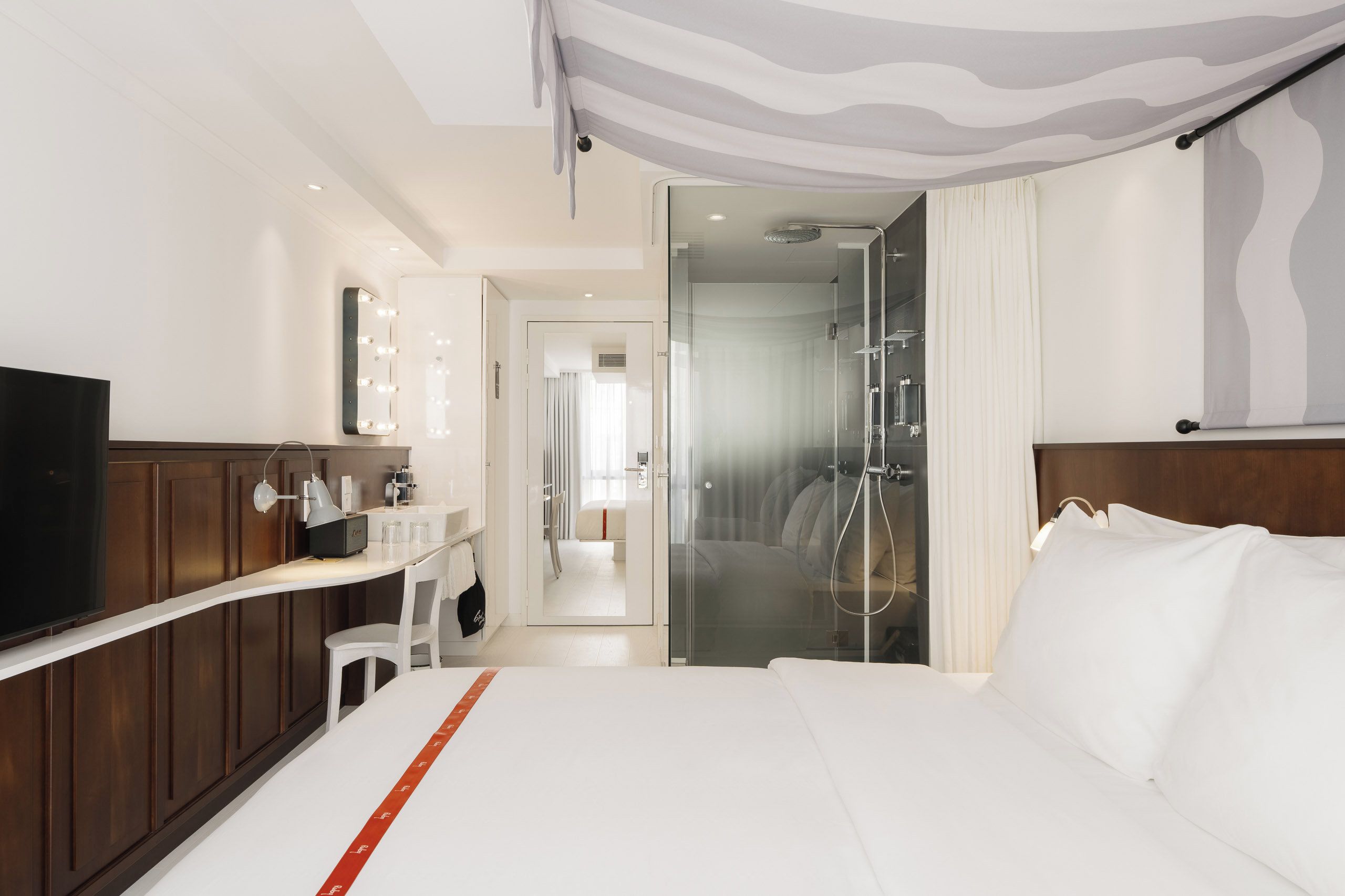 A hotel room with a bed, a TV, a vanity with a mirror, a shower, and a hallway leading to another room.