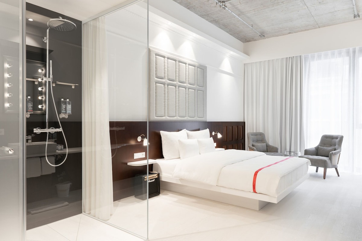Une chambre d’hôtel moderne dotée d’une douche en verre, d’un grand lit avec une literie blanche, d’une tête de lit au motif capitonné et de deux fauteuils.