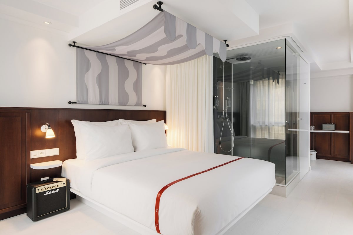 Ein modernes Hotelzimmer mit einem großen Bett, einer gläsernen Dusche, einer Minibar und einer Marshall-Stereoanlage.