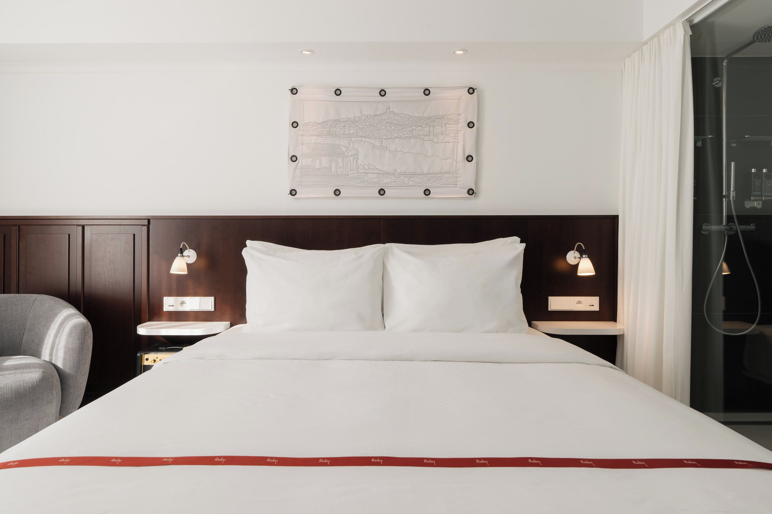 A hotel room with a large bed, two bedside tables, a chair, a wall-mounted artwork, and a shower area.