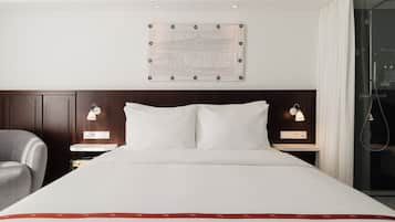 A hotel room with a large bed, two bedside tables, a chair, a wall-mounted artwork, and a shower area.
