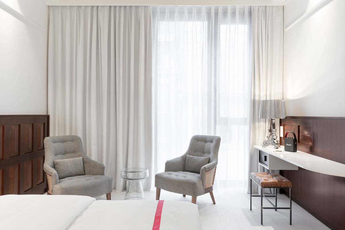 Ein Hotelzimmer mit einem Bett, zwei Sesseln, einem kleinen Tisch, einem Schreibtisch mit Lampe und einem großen Fenster mit Vorhängen.