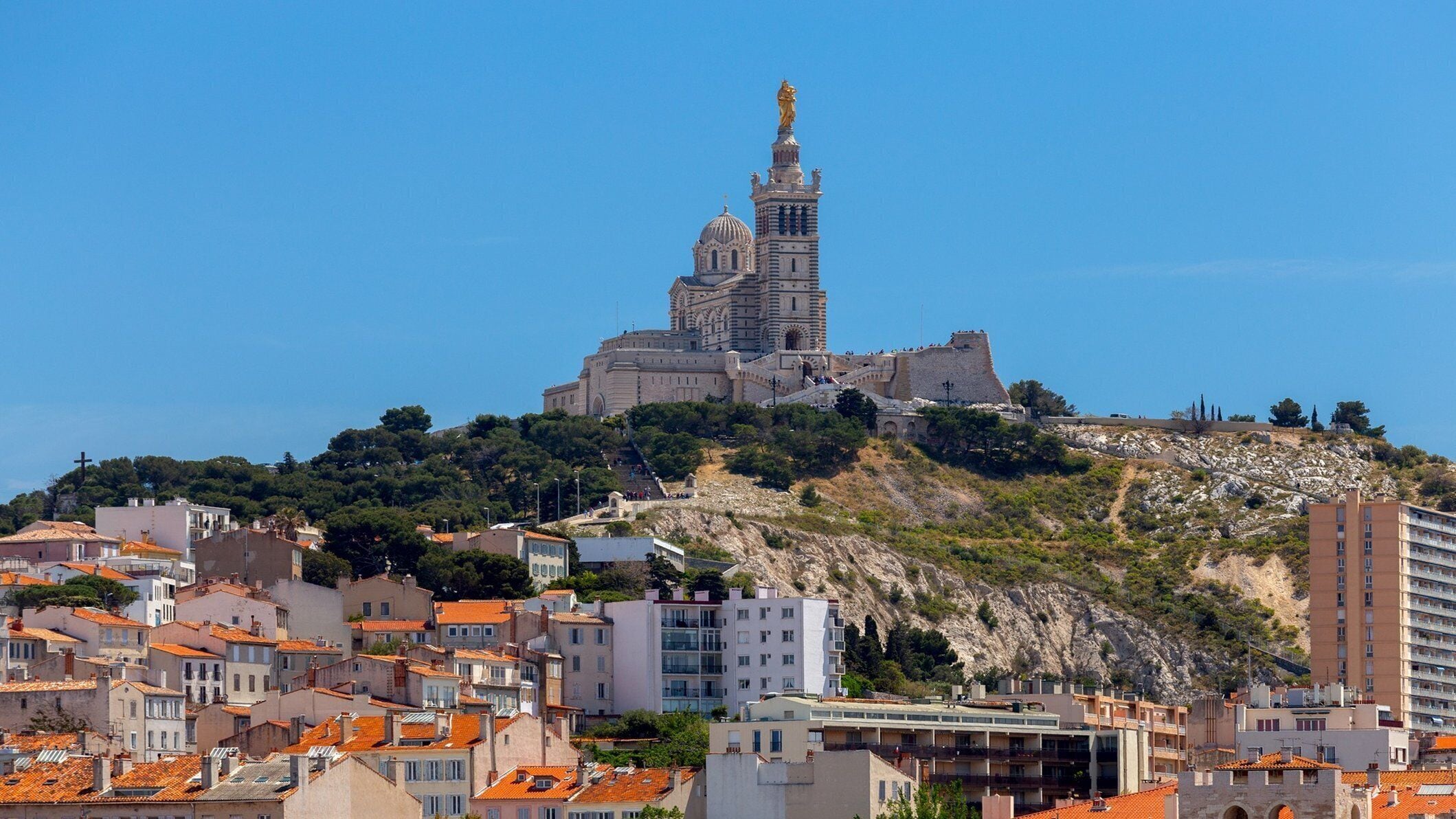 Un bâtiment historique doté d’une tour imposante et d’une statue, entouré d’immeubles résidentiels aux toits recouverts de tuiles rouges.