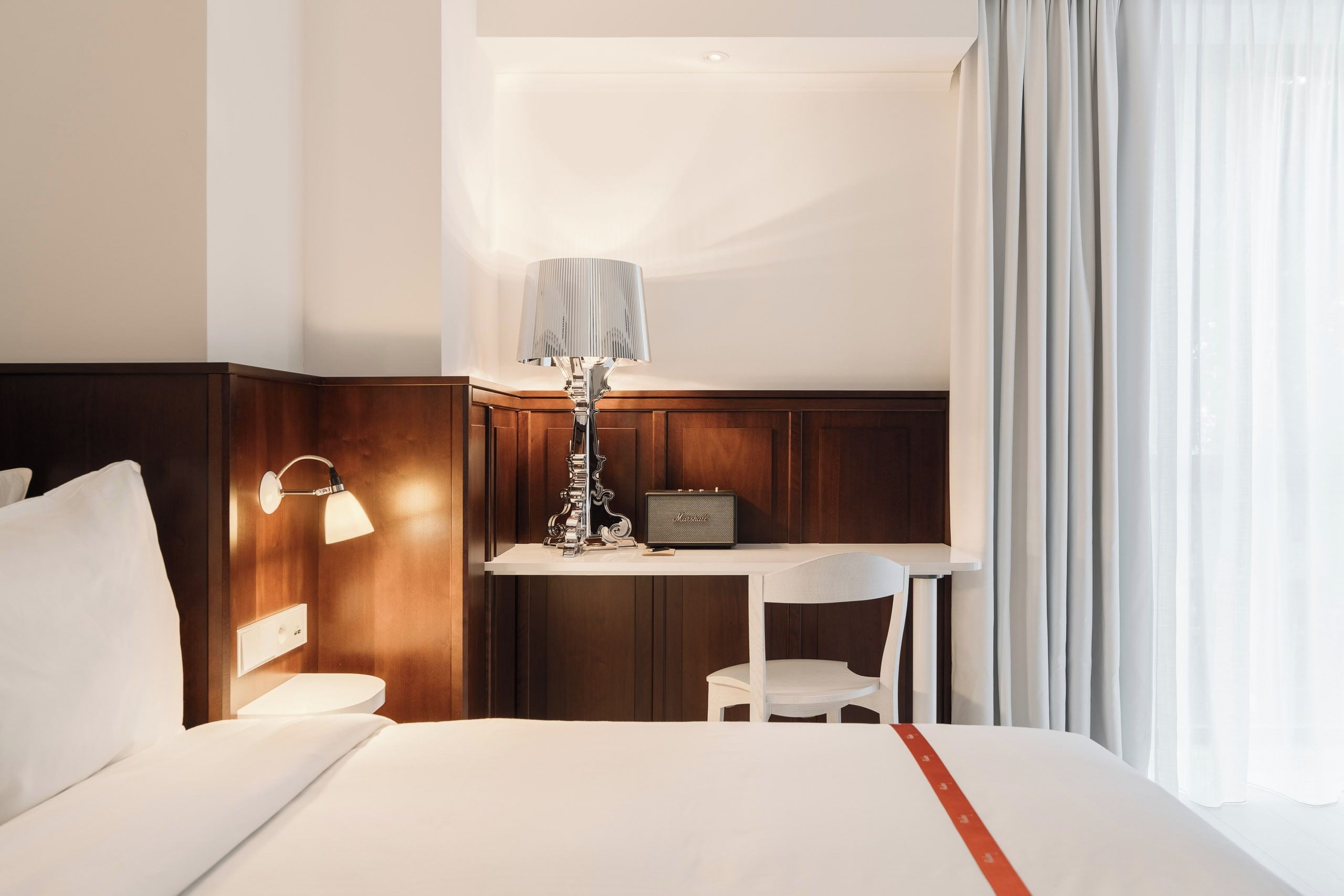 A hotel room with a bed, a desk, a lamp, a chair, and a window with curtains.