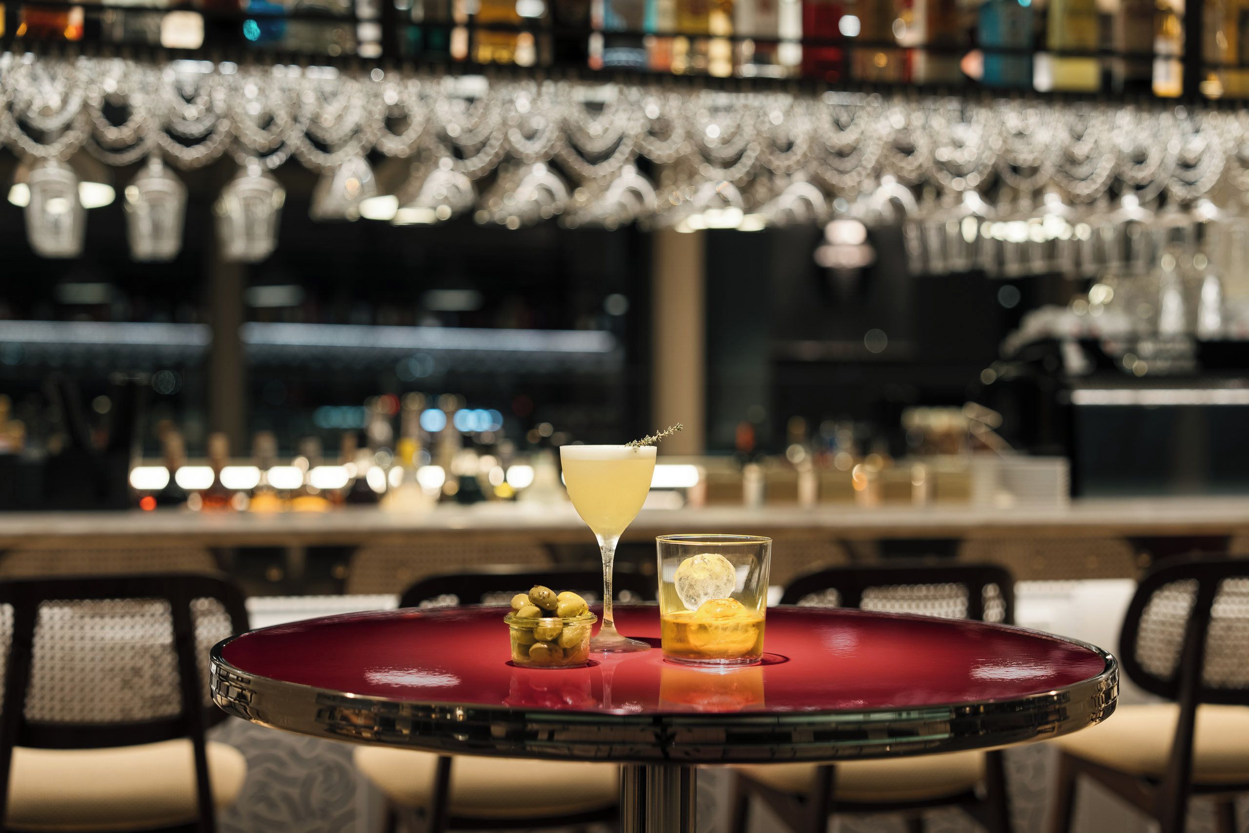 A bar setting with a red table, a cocktail glass, olives, and a shot glass with an ice cube.