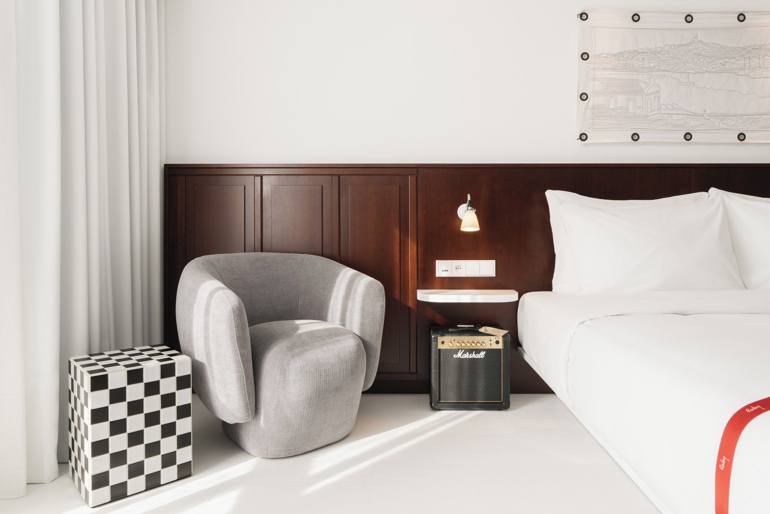 Une chambre d’hôtel avec un lit, un fauteuil gris, une petite table avec une lampe, un amplificateur Marshall et un cube à motifs à carreaux.