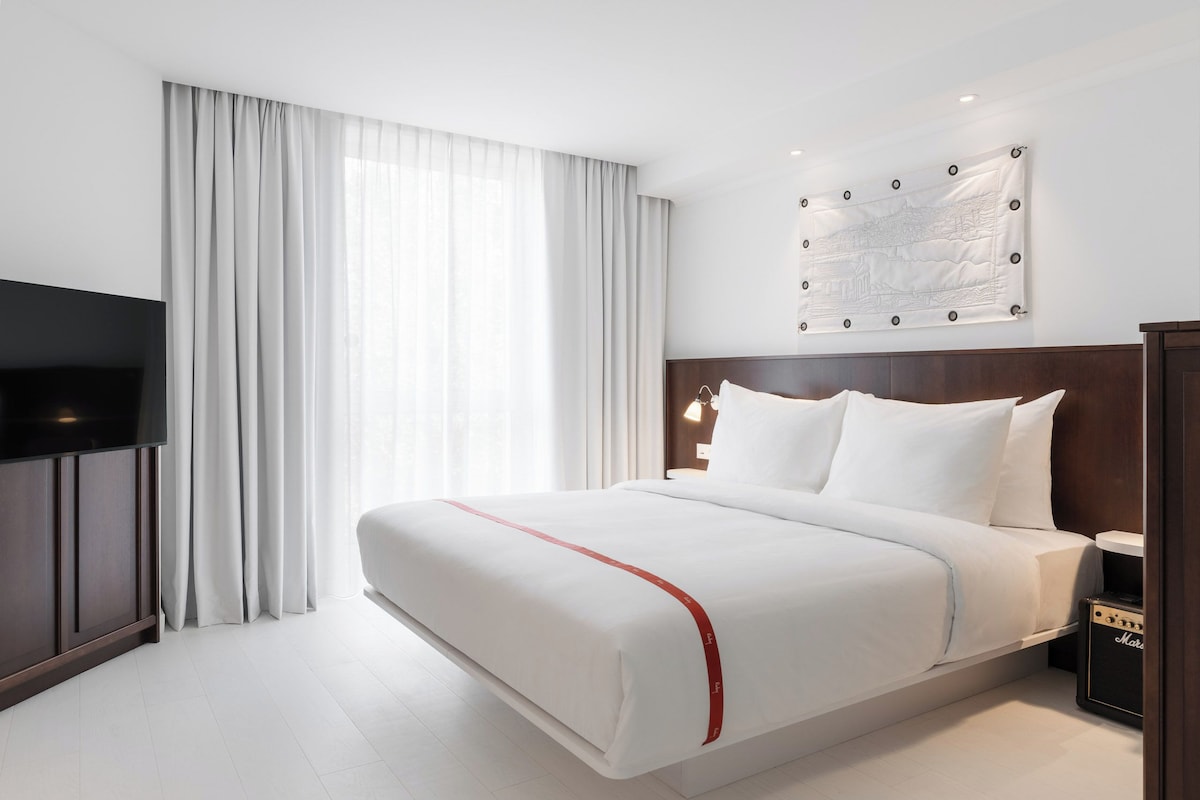 Ein Hotelzimmer mit einem großen Bett, einem Flachbildfernseher, einem Wandbild und einer roten Bettdecke mit Streifen.