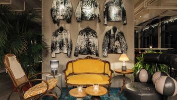 Un hall d’entrée avec un canapé jaune, un fauteuil en osier, un fauteuil en cuir noir, une petite table ronde avec une lampe, et un mur où sont accrochées plusieurs vestes.