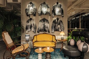 A lobby with a yellow sofa, a wicker chair, a black leather chair, a small round table with a lamp, and a wall with several jackets. - Ruby Lilou Hotel Marseille by IHG (Marseille)