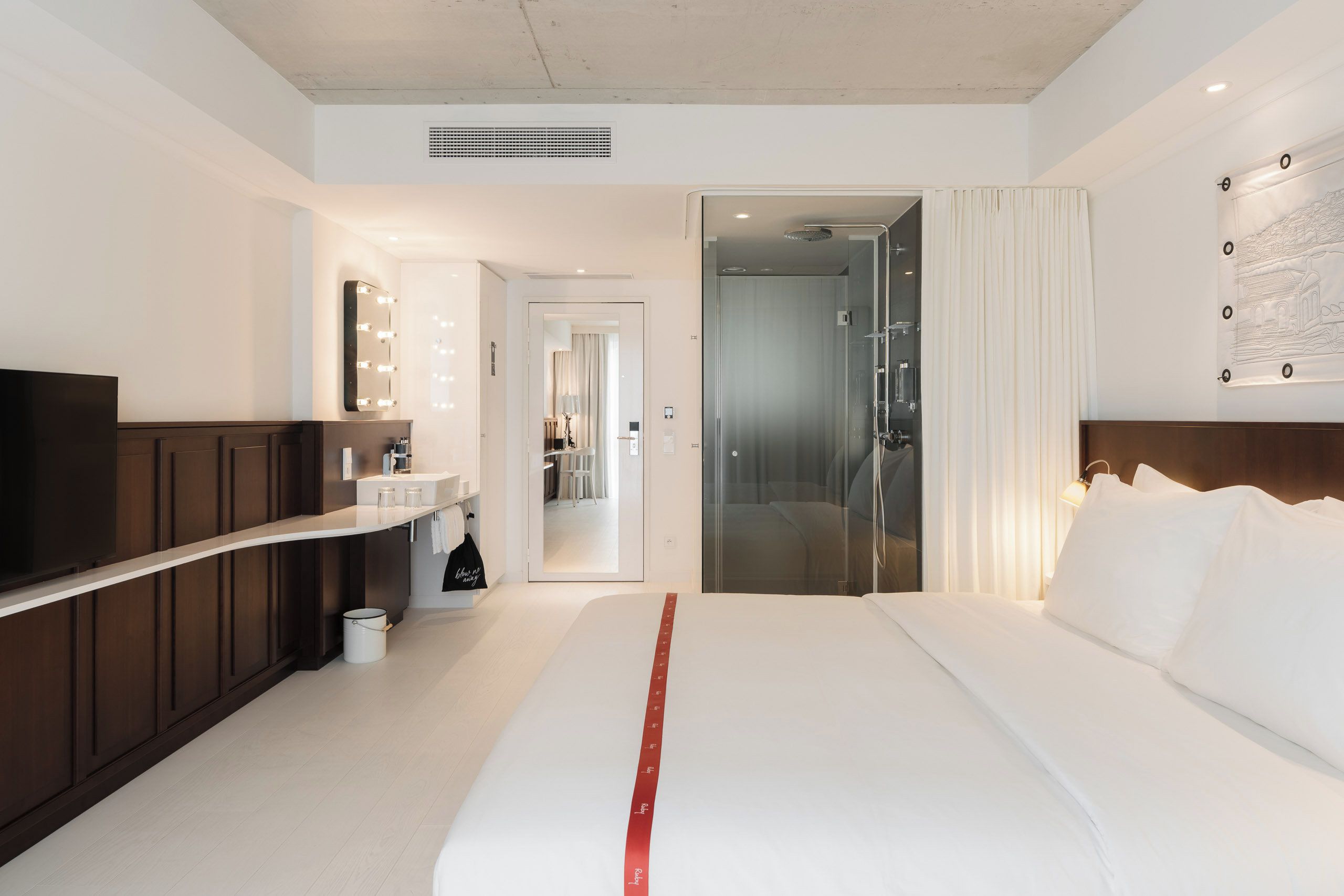 A hotel room with a large bed, a TV, a bathroom with a shower, and a hallway leading to another room.