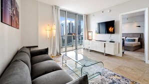 Una sala de estar moderna con un sofá, una mesa de centro de vidrio, un televisor y vistas a la ciudad a través de amplios ventanales.