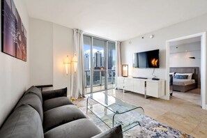 Ein modernes Wohnzimmer mit einer Couch, einem Glastisch, einem Fernseher und Blick auf die Stadt durch große Fenster.