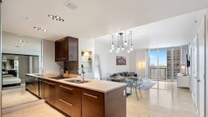 Una cocina moderna con una isla central, un comedor con mesa de vidrio y vistas a un edificio de gran altura.