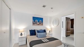 Ein Schlafzimmer mit einem Bett, Nachttischen, einem Gemälde, einem Spiegel und einer Tür, die in eine Küche führt.