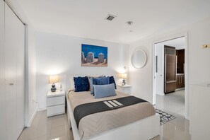 Ein Schlafzimmer mit einem Bett, Nachttischen, einem Gemälde, einem Spiegel und einer Tür, die in eine Küche führt.