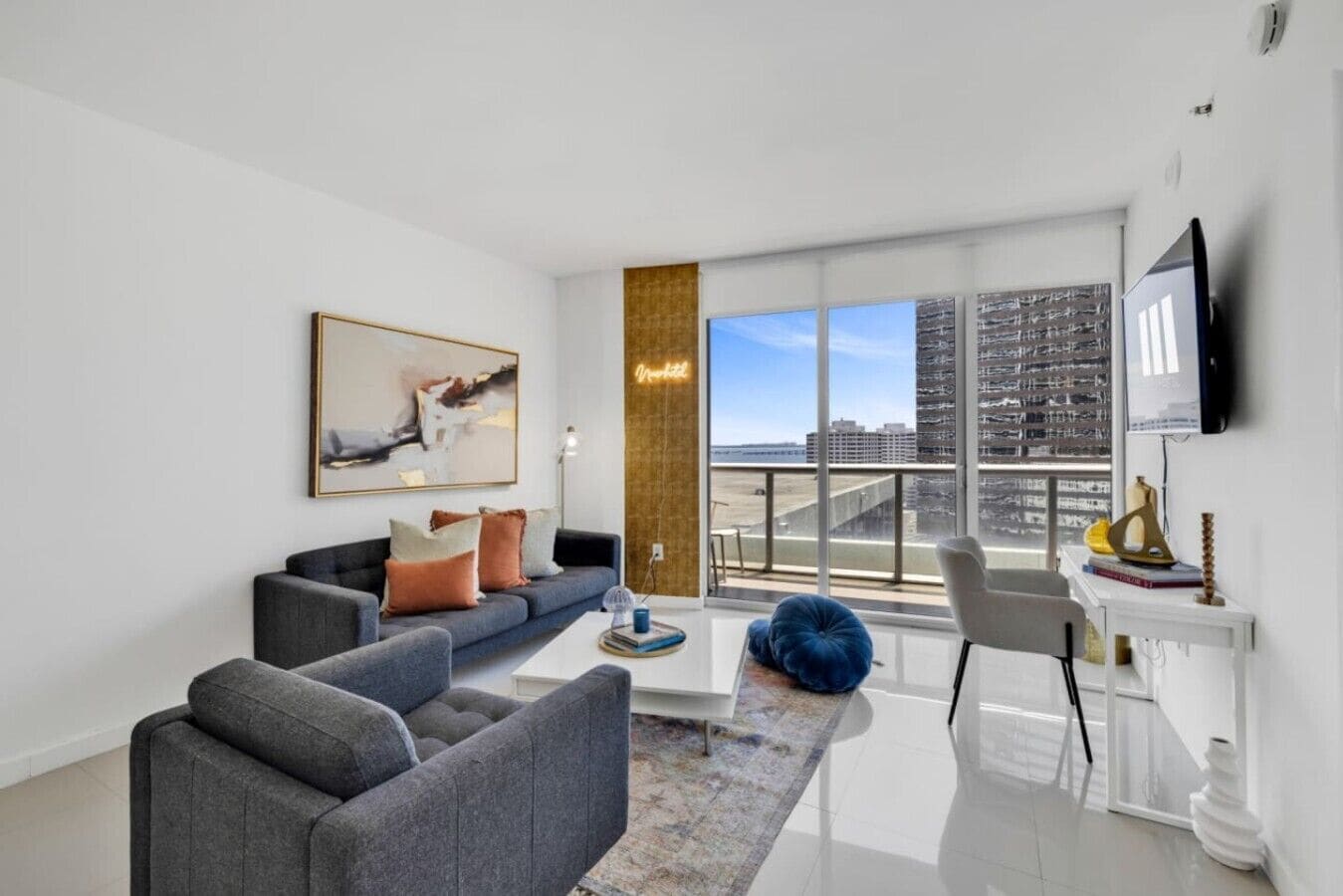 A modern living room with a grey sofa, a coffee table, a blue bean bag, a painting, and a city view through large windows.