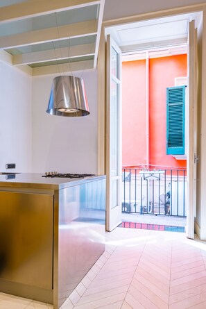 A modern kitchen with a stainless steel island, a hanging pendant light, and a view of a balcony with a red wall and blue shutters. - Chiaia Prestige by NapoliApartments (Naples)
