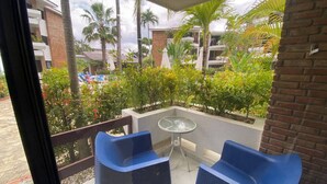 Een balkon met twee blauwe stoelen en een glazen tafel, met uitzicht op een tuin met palmbomen en een gebouw op de achtergrond.
