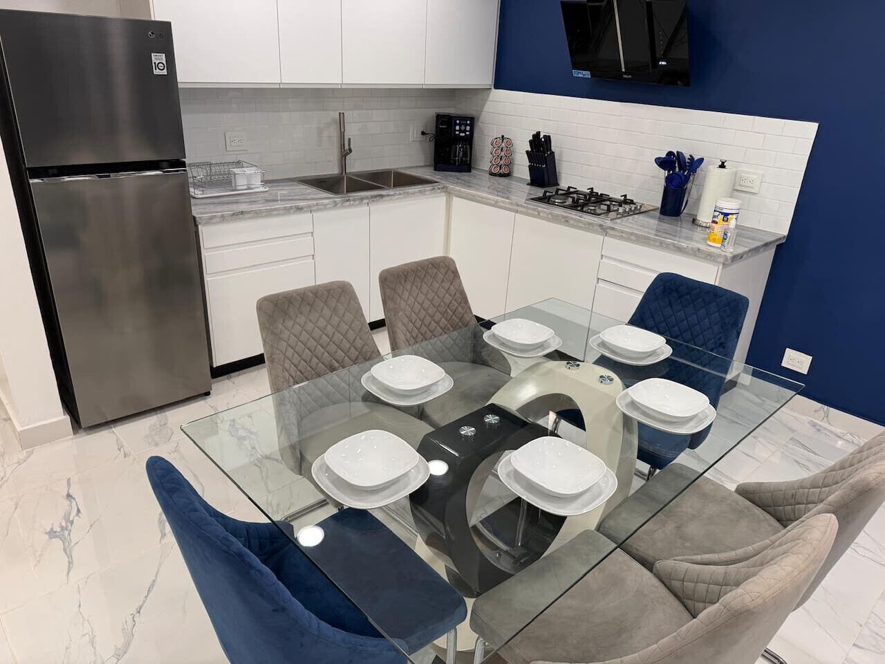 A modern kitchen with a glass dining table, white plates, and blue chairs.