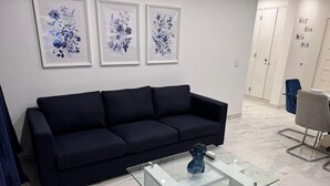Een moderne woonkamer met een donkere bank, een glazen salontafel en bloemachtige kunstwerken in lijsten aan de muur.