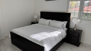 Een slaapkamer met een groot bed, nachtkastje, lamp en raam met uitzicht op gebouwen.