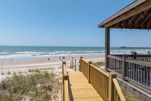 Ein hölzerner Steg führt zu einem Strand, in der Ferne ist ein Pier zu sehen.