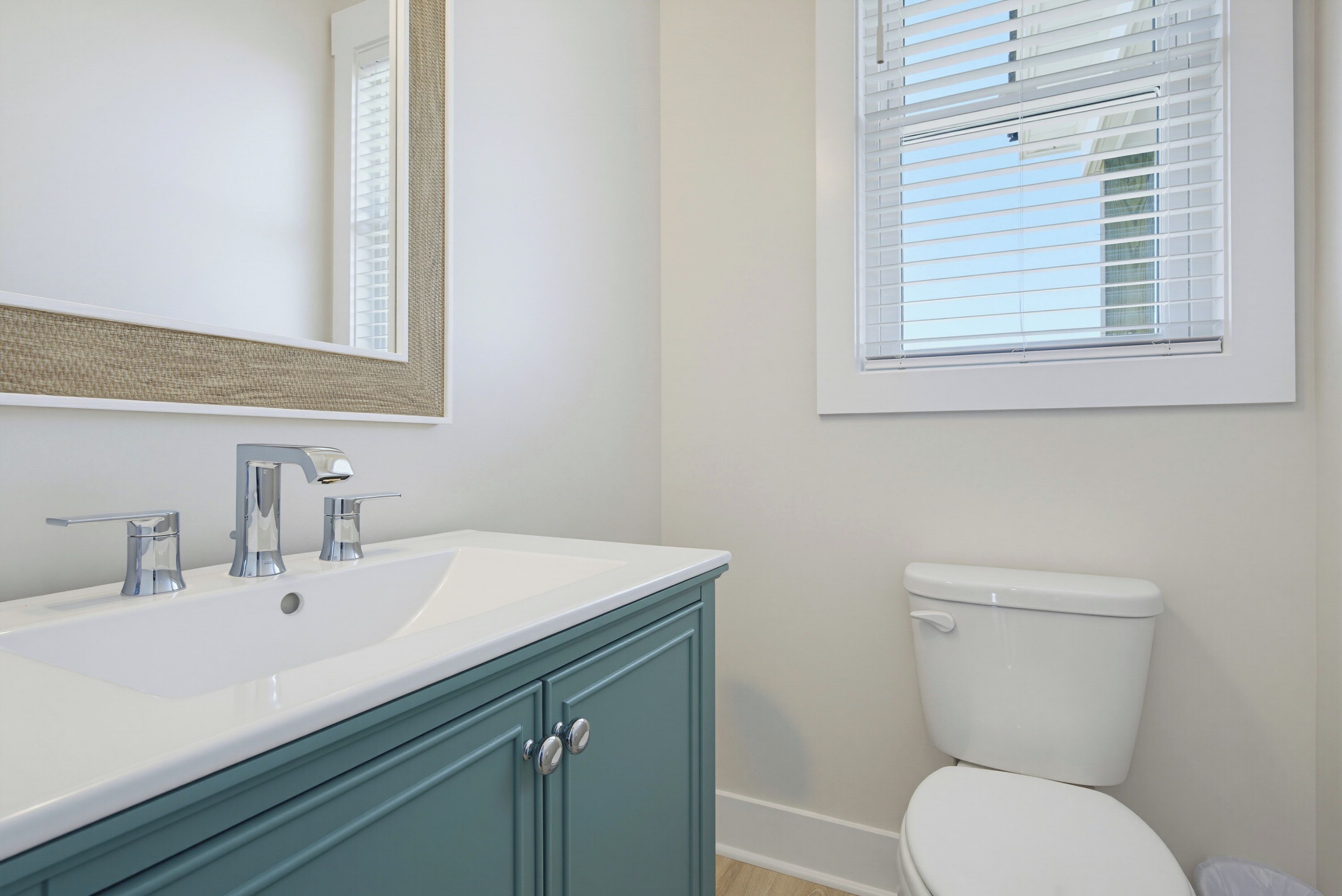 Un baño con lavabo blanco, espejo, inodoro y ventana con persianas.