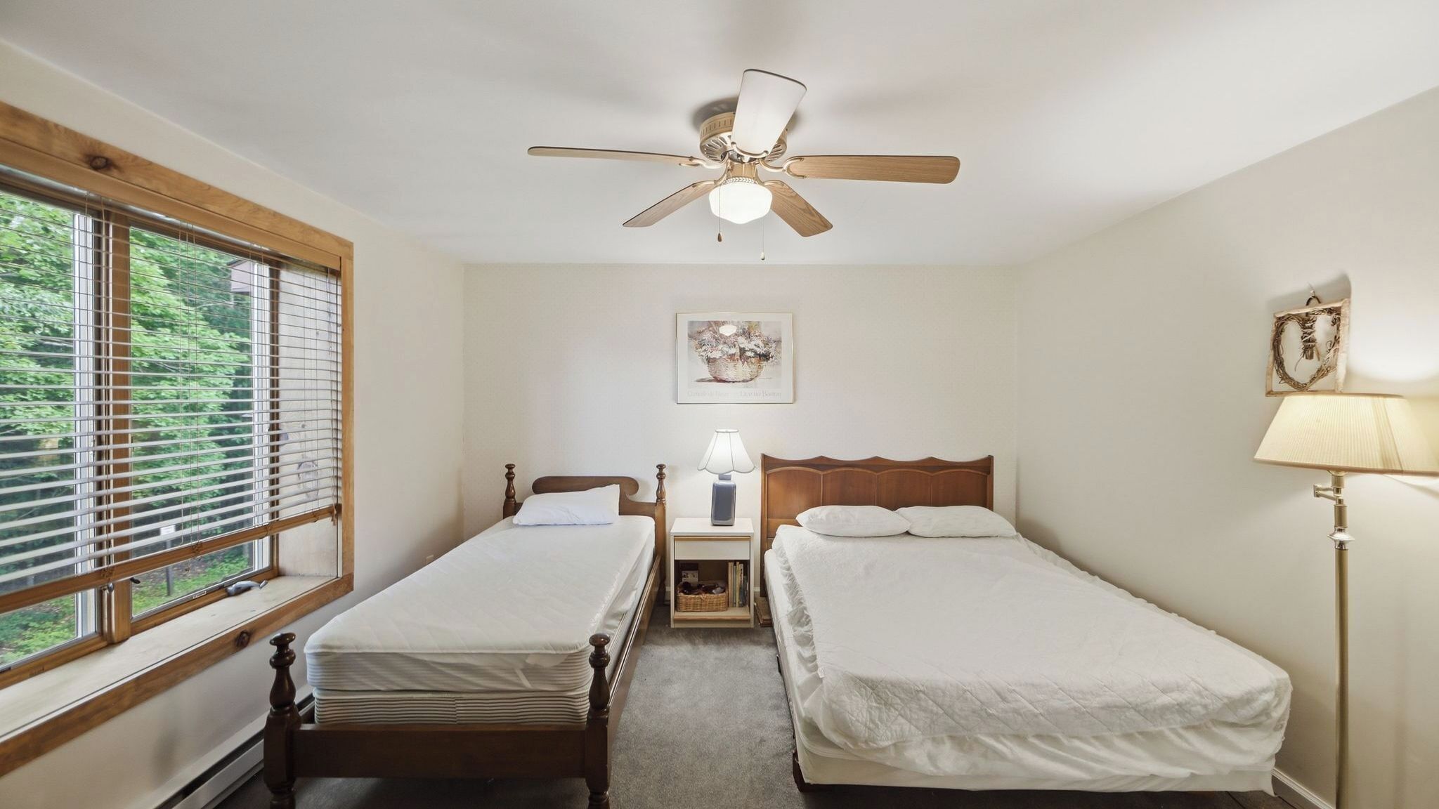 Un dormitorio con dos camas individuales, un ventilador de techo, una mesita de noche con lámpara, un cuadro en la pared y una ventana con persianas.