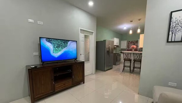 Ein Wohnzimmer mit Fernseher an einer Holzvitrine, einem Essbereich und einer Küche im Hintergrund, sowie einem Flur, der in einen weiteren Raum führt.