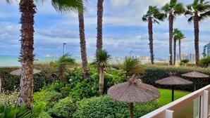 Een aangelegde tuin met palmbomen en rieten parasols, een balkon met uitzicht op een waterpartij en een verafgelegen stadsbeeld.