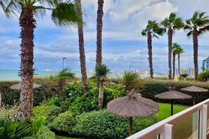 Ein gepflegter Garten mit Palmen und Reetdachschirmen, ein Balkon mit Blick auf ein Gewässer und eine entfernte Skyline.