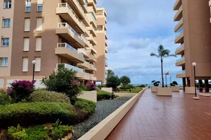 Ein mehrstöckiges Hotel mit Balkonen, einem gepflasterten Weg und begrünten Außenanlagen mit Sträuchern und Bäumen.