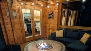 Interior de madera con una chimenea redonda, asientos azules y una puerta de cristal que da a otra habitación.