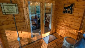 Interior de una cabaña de madera con puerta de vidrio, una cama con almohadas, un adorno de oso y un cartel que dice 'HOME SWEET HOME'.