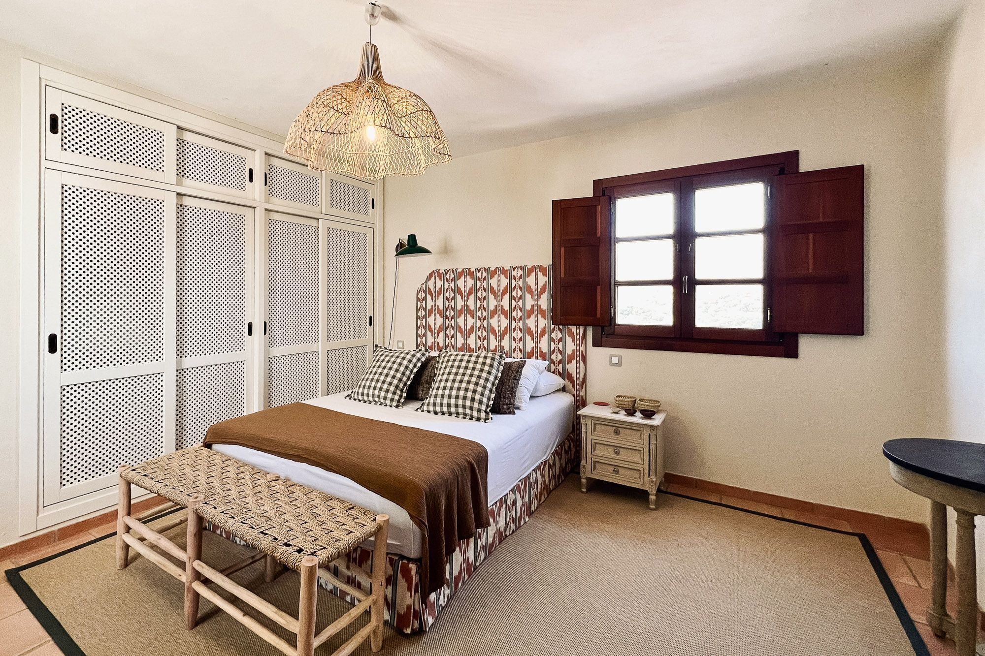 Un dormitorio con cama, mesita de noche, lámpara colgante, ventana con contraventanas y armario con diseño perforado.
