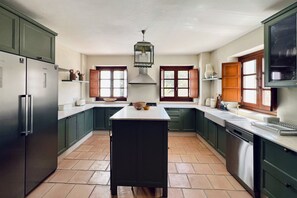 Eine Küche mit grünen Schränken, einer zentralen Kochinsel, einem Edelstahlkühlschrank und einem Fenster mit Holzjalousien.
