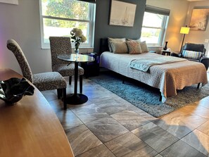 Um quarto com cama, uma cadeira, uma mesinha e um vaso com flores.