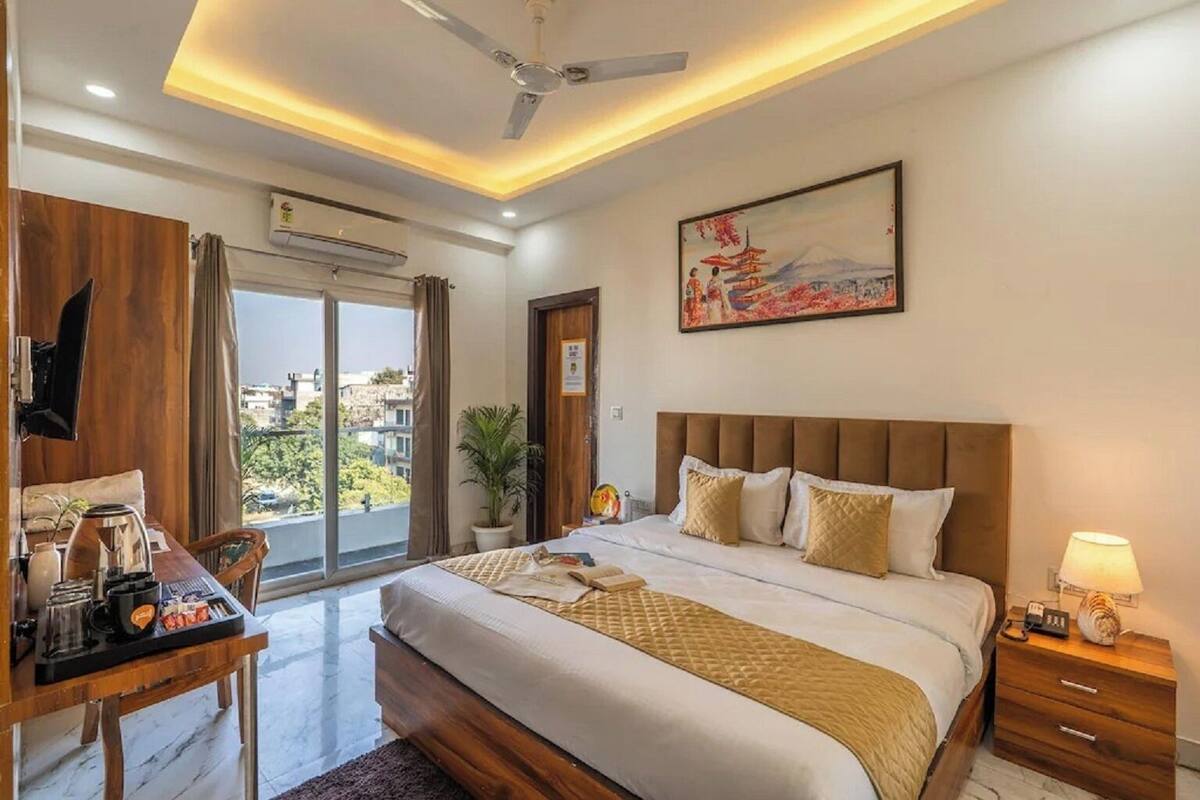 A hotel room with a bed, bedside table, desk with coffee maker, and a painting on the wall.