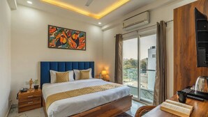 A hotel room with a bed, a desk, a chair, a painting, a window, and an air conditioning unit. - Coco House (Greater Noida)