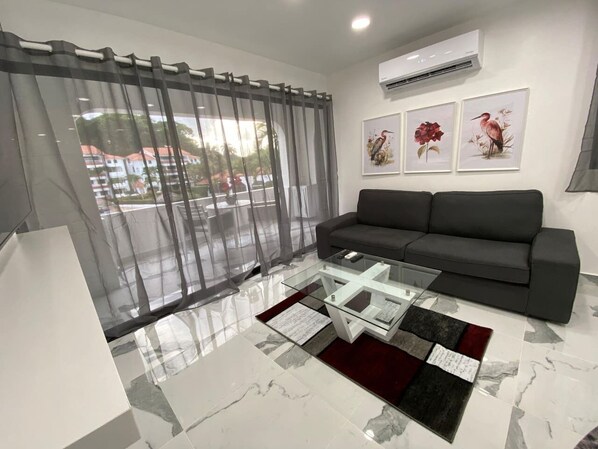 Een moderne woonkamer met een glazen salontafel, een grijze bank en een groot raam met dunne gordijnen.