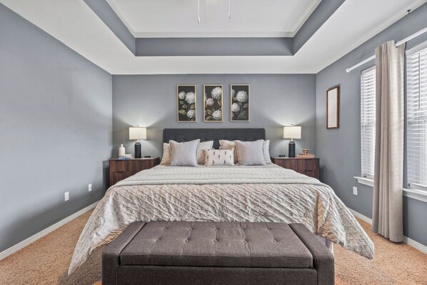 A bedroom with a bed, two bedside tables, a bench, a window with curtains, and wall art.