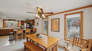 A kitchen with wooden cabinets, a dining table with chairs, a ceiling fan, and a window with a view of houses. - Lower Darby by Avantstay Indoor Heated Pool + Billiards + Foosball Near Gore (North Creek)