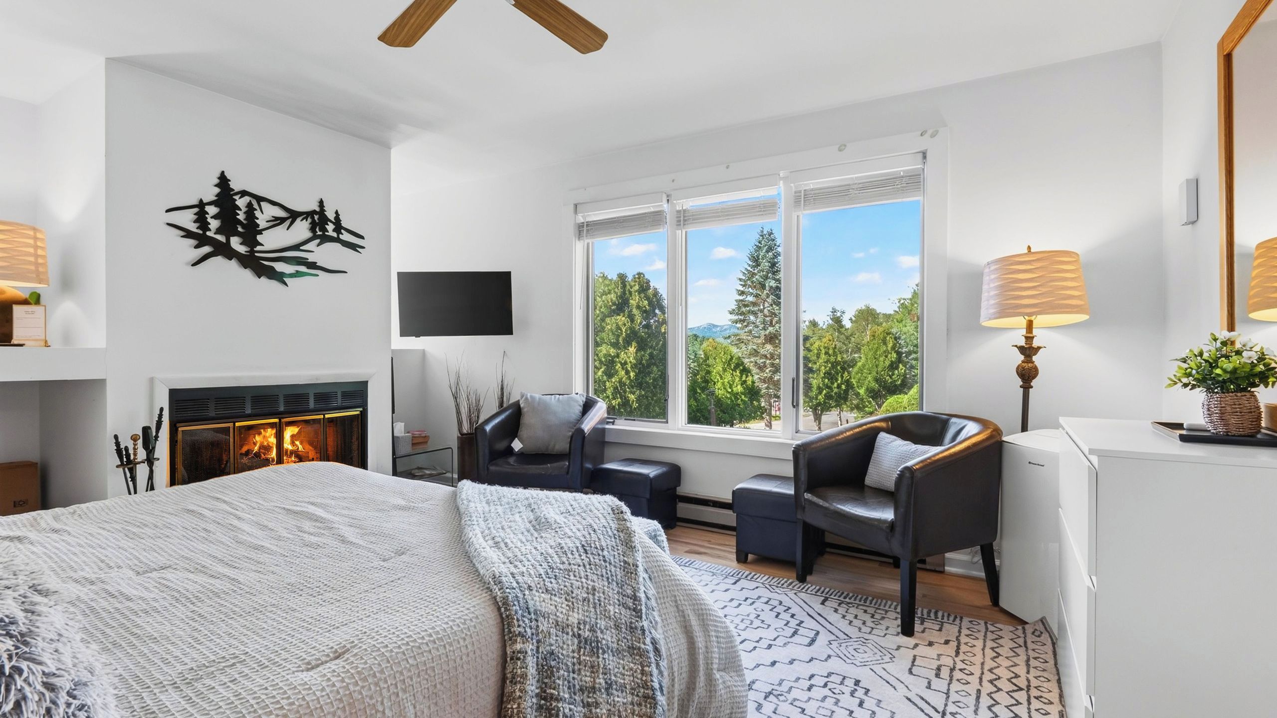 A modern bedroom with a fireplace, a large bed, a seating area with a view of the outdoors, and a television on the wall.